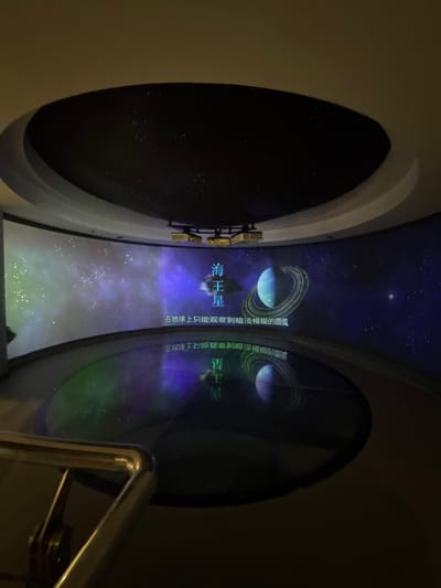 The image shows a planetarium-like view through a circular opening. The celestial display features a planet with prominent rings, resembling Saturn or Uranus, surrounded by stars and cosmic dust. Text in Chinese is displayed, referring to Neptune (海王星) and mentioning that only a faint and blurry arc can be observed from Earth. A nearby circular window reflects a reversed image of the celestial display and the text. A metal railing is visible on the right side.
