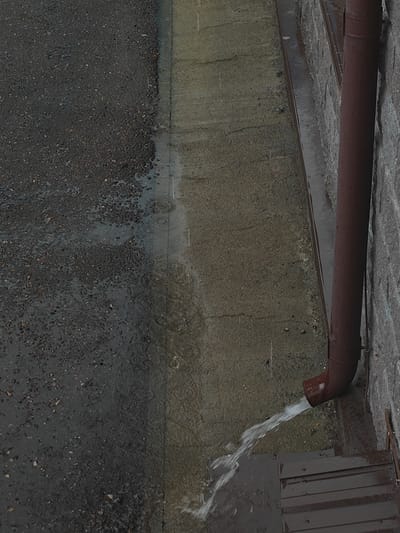 In Дүңгілағаш, Kazakhstan, this image depicts a close-up, high-angle view of a downspout discharging a stream of water onto a metal surface, likely a set of stairs or a grate, adjacent to a textured concrete wall. The downspout is a reddish-brown cylindrical pipe that curves downwards. The surrounding ground is composed of dark asphalt with small gravel, showing numerous wet patches and ripples indicative of rainfall. A narrow strip of lighter-colored, wet concrete runs alongside the asphalt, bordered by a dark metal strip. The overall scene suggests an outdoor urban environment during or immediately after a rainfall. No people are present in the image.