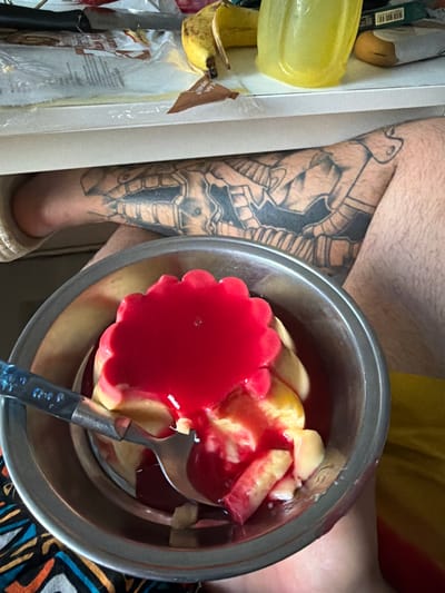 This image shows a close-up of a person's tattooed leg and a bowl of dessert. The tattoo appears to be a black and grey design with intricate details, possibly depicting mechanical or armored elements. The dessert in the metal bowl consists of several pieces of what looks like pudding or custard, topped with a vibrant red jelly or syrup. A spoon with a dark handle is resting in the bowl, suggesting someone is about to eat or has been eating the dessert.
In the background, to the left of the person's leg, there are some kitchen items. A bright yellow plastic container is visible, along with what appears to be a peeled banana and some packaging materials, possibly for food. A white vertical surface, likely a cabinet or wall, forms a boundary on the left side of the image. The overall lighting suggests it is daytime, but the specific time is not discernible. There are no clear indicators of the weather or the exact location beyond the provided context of Boxtel, Netherlands. The focus is on the contrast between the detailed tattoo and the appetizing dessert.