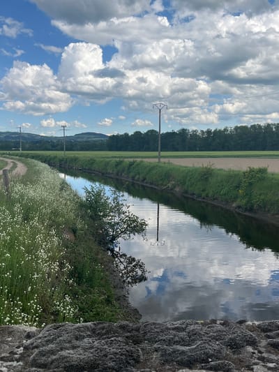 The image captures a rural scene near Saint-Just-Saint-Rambert, France. A narrow canal or stream runs horizontally across the middle of the image, reflecting the cloudy sky above. The waterway is bordered on both sides by grassy banks. One bank features white wildflowers in bloom. Power lines run along the left side, supported by utility poles. Beyond the canal, fields and trees stretch into the distance, with a slight hill visible on the horizon. A rough rock wall is visible on the right side of the image. The sky is filled with puffy white clouds against a blue backdrop.