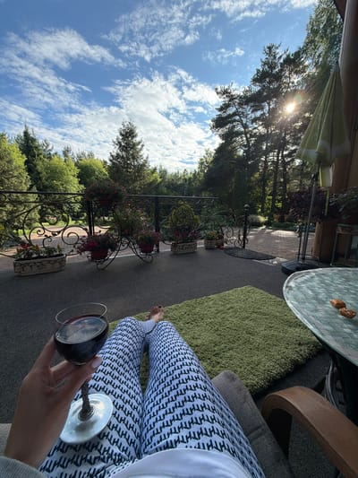 The image shows a first-person perspective of a person relaxing on a balcony or patio, holding a glass of red wine. The person is wearing patterned leggings and has painted toenails. In the foreground, a green rug is laid out on the floor. To the right, a glass-topped table with two nuts on it is visible, along with a chair. Behind the table, an umbrella is partially visible. The background features a wrought-iron railing with potted plants and flowers. Beyond the railing, a paved area leads to a lush green garden with mature trees and foliage under a bright, partly cloudy sky. The sun is visible, casting light through the trees. The scene suggests a peaceful outdoor setting, likely at a private residence in Nevzorovo, Russia, given the context of the request.