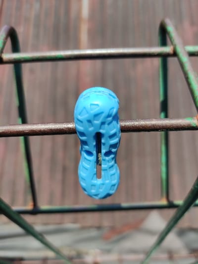 The image displays a single, light blue plastic clothespin, distinctly shaped like a peanut with a textured surface, clipped onto a horizontal, rusted metal bar. The clothespin features a prominent central vertical slot, within which faint green-brown residue is visible. This metal bar is part of a larger, weathered metal framework exhibiting significant rust and traces of faded green paint. The background is blurred, showing predominantly reddish-brown vertical wooden planks or siding. The scene is illuminated by bright, likely natural, light. The documented location of this scene is Staufenberg, Germany.