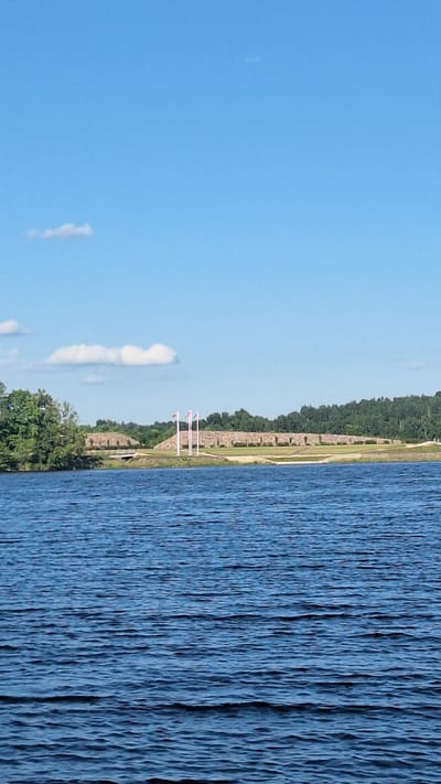 The image shows a wide body of rippling blue water in the foreground, stretching towards a distant shore. On the shore, a stone-built structure resembling a fortification or embankment dominates the mid-ground. Three flagpoles stand prominently in front of this structure, each flying a white flag with a red cross. Beyond the fortification, a dense line of dark green trees forms a backdrop, suggesting a natural landscape. The sky above is a clear, bright blue with a few wispy white clouds scattered across it. The overall scene appears to be a calm, outdoor environment, possibly a park or historical site near a body of water. The location is identified as the historic fort of Vecpiebalga in Sece, Latvia.