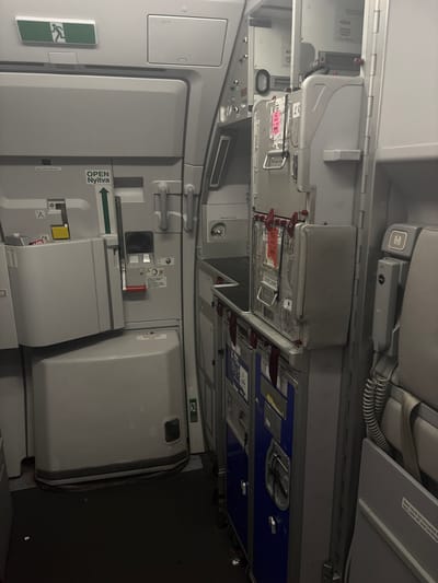 The image depicts the interior of an aircraft, specifically an aisle area with a galley cart. On the left, a large, grey, partially opened door is visible, with an illuminated green exit sign above it. Below the exit sign, there's a sign that reads "OPEN NYITVA" with an upward-pointing arrow. Further down the door, a red handle is present.
To the right of this door, a metal galley cart is positioned, filled with various compartments and latches. Some labels and stickers are affixed to the cart. Above the galley cart, there are panels and storage compartments within the aircraft's structure. On the far right, airplane seats are partially visible, along with a gray telephone handset mounted on the wall. The floor is covered with a dark, textured carpet.
The image does not provide any information about the city of Pitești, Romania, nor does it contain any contextual clues to suggest this location. It is a forensic, matter-of-fact description of the interior of an aircraft.