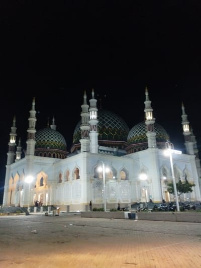The image presents a large mosque in Kotapantonlabu, Indonesia, photographed at night under artificial illumination. The sky is completely dark and featureless. The mosque exhibits a prominent white facade with intricate architectural details.
At least three large domes are visible, each adorned with a distinct green, white, and golden or yellow patterned surface, specifically displaying a diamond or grid-like motif on the green areas. The building is flanked by at least six tall, slender minarets, also white, topped with pointed finials, and featuring internally lit sections in their upper tiers. The mosque's lower walls incorporate multiple arched doorways and windows, with some sections detailed by intricate white decorative patterns.
The foreground consists of a paved area, transitioning from a light-colored concrete or asphalt surface to a reddish-brown brick-patterned pavement. Scattered debris is visible on the lighter paved section. Artificial streetlights, positioned in the foreground and nearer the mosque, cast bright, yellowish-white light on the ground and parts of the building. A low concrete barrier separates the immediate foreground from the mosque's grounds. Several indistinct human figures are visible near the mosque's entrances and steps, some standing, others seated. A cluster of motorcycles is parked behind the concrete barrier on the right side of the image, adjacent to a small tree.