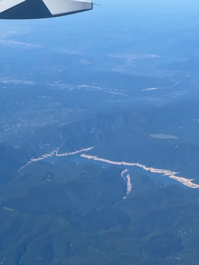This is an aerial photograph taken from an airplane, looking down at a landscape characterized by mountains covered in dark green forests and a body of water that appears to be a lake or reservoir. The water's edge is irregular, with exposed, lighter-colored soil or rock formations suggesting a low water level. The mountainous terrain is densely forested, with visible valleys and slopes. In the far distance, a faint haze obscures the details of the land, but hints of human settlement, possibly a town or city, can be discerned. The tip of an airplane wing is visible in the upper left corner of the frame, indicating the perspective of the photograph. The question asks to include the location in the city of Mogoșoaia, Romania; however, based on the visual information provided in the image, it is not possible to confirm or identify any specific landmarks or features that would definitively place the scene in Mogoșoaia or any other specific city or location. The image depicts a natural landscape with signs of distant civilization, but lacks the specific detail required for precise geographical identification.