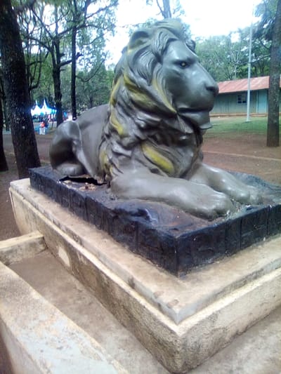 The image displays an outdoor scene in the city of West Sakwa ward, Kenya, featuring a weathered statue of a recumbent lion. The lion, dark grey-green with faded yellow highlights on its mane and prominent yellow eyes, faces right. It rests on a dark, textured rectangular base, which is elevated by a multi-tiered light grey concrete pedestal. A visible crack is present on the lower left flank of the lion statue, near its base. In the background to the left, tall trees with dark trunks and green foliage partially obscure a blue and white tent structure, near which several indistinct human figures are visible. To the right, a reddish-brown dirt path leads to a light blue building with a rusty red corrugated metal roof and a thin vertical pole in front of it. Additional trees are visible in the distant background. The scene appears to be an open, park-like area. No direct human actions or interactions are depicted in the immediate foreground.