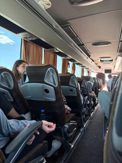 The scene is inside a passenger bus. Rows of seats are visible, most of which are occupied by people. Several passengers are visible, including one wearing a white shirt, and another in a grey sweatshirt. A water bottle is visible in a seat-back pocket. The bus windows show a clear blue sky. Location information (Yazdach, Bulgaria) is not discernible from the image.