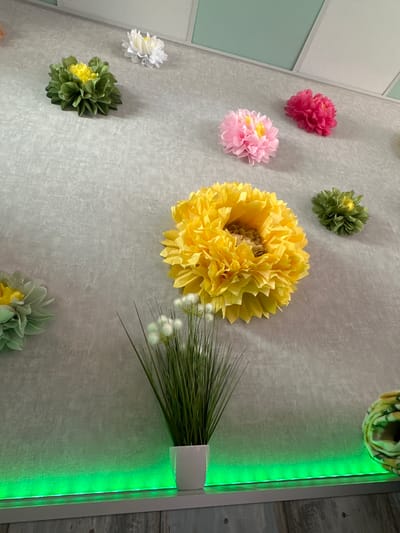 This media file shows a wall decorated with various artificial flowers. The flowers are of different colors, including pink, yellow, green, and white. They appear to be made of tissue paper or a similar material and are arranged in a scattered pattern on a textured gray wall. In the upper left corner, there are some geometric wall panels, one of which is light blue-green and the others are white. To the right, there is a white vertical frame containing a strip of green LED lights, illuminating the wall with a bright glow. A bunch of green faux grass is placed in a white holder attached to this frame, with some small white flower accents. The overall impression is of a decorative wall display, possibly in a room. There is no visible text in the image.
