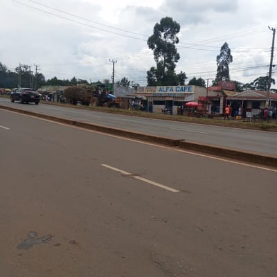 The image depicts a street scene in Sheywe ward, Kenya. A car is traveling on a two-lane road with a concrete median. On the right side of the road are several small businesses, including Alfa Cafe and a butcher shop. There are people walking and standing in front of the shops. A tractor carrying a load of material is also visible on the right side of the road. Overhead power lines are strung across the scene. The sky is cloudy.