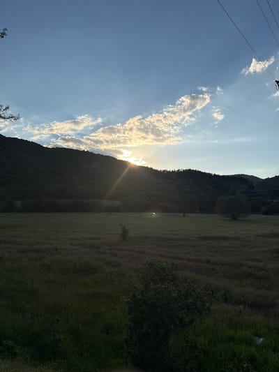 The image shows a landscape at Mesta, Bulgaria. The scene includes a mountain range silhouetted against a sky filled with bright clouds and sunlight. A field extends to the right of the mountain, with varying shades of green suggesting different vegetation. A powerline is visible in the upper left corner.