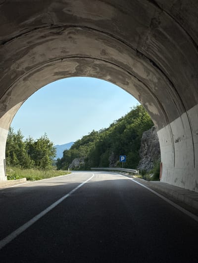 The media file shows a view from inside a concrete tunnel, looking out towards a bright, clear sky and a lush green mountainside. The tunnel opening is circular and imposing, with rough concrete textures and visible lines of construction. On the right side of the frame, a paved road curves away, marked with a white line. Beyond the road, the mountainside is covered in dense green trees, with a rocky cliff face visible higher up. A blue parking sign with a white "P" is mounted on a pole beside the road, indicating a possible resting or viewing area. The time of day appears to be midday, with strong sunlight casting shadows. The weather is clear and fair. The overall scene suggests a scenic route through mountainous terrain, likely in a rural or natural setting. The image evokes a sense of transition, moving from the dark, enclosed space of the tunnel to the open expanse of nature. There is no visible text other than the letter "P" on the parking sign. The location is identified as Dragalj, Montenegro.