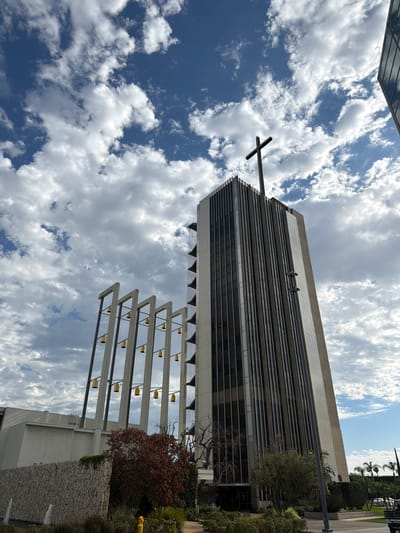 This is a low-angle, full exterior, eye-level shot of Christ Cathedral in Garden Grove, California, United States of America. The church, formerly the Crystal Cathedral, features a tall, modern tower with a cross at its apex. The tower is flanked by a structure with vertical lines and a series of golden bells suspended from angled supports. The building is made of concrete and glass. In the foreground, a stone wall runs horizontally across the frame, with a small fountain and some vegetation. The sky is blue with scattered white clouds. A person is visible on a distant bench.