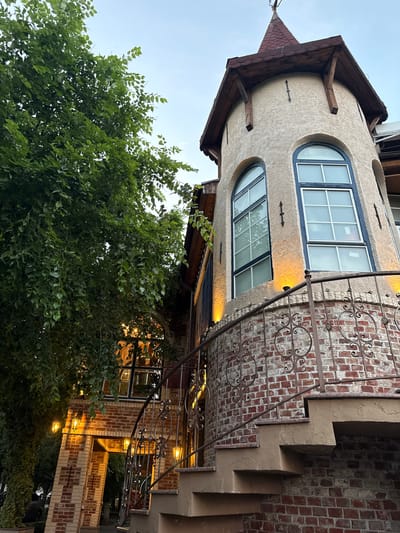 The image shows an exterior view of a building with a unique architectural style, likely located in Oktyabr'skoe, Russia. The building features a stucco facade with arched windows and a reddish-brown brick exterior on a staircase and a section of the wall. Decorative metal railings with ornate scrollwork are attached to the bricked areas, and outdoor lights are visible, suggesting it is dusk or nighttime. Lush green foliage from a tree partially obscures the lower portion of the building. The overall atmosphere appears somewhat whimsical and charming, with the warm glow of the lights contrasting with the cool tones of the sky. There is no visible text in the image.
