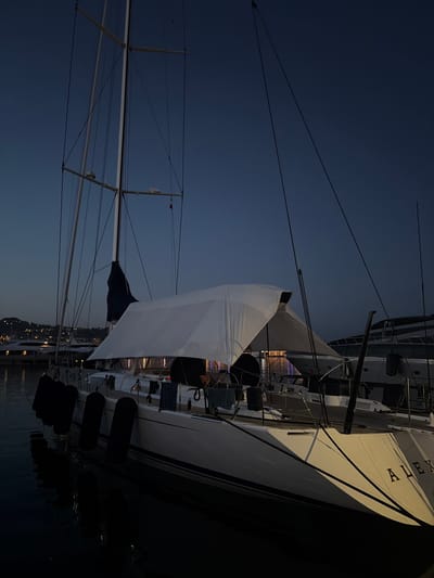 A sailboat named "ALEX" is docked at night in the city of Imperia, Italy. The sailboat has a white hull and a dark blue sail furled on the mast. A white tarp is stretched over the boom, creating a makeshift shelter on the deck. The boat is equipped with several black fenders hanging along its side. In the background, other boats and distant city lights are visible. The water appears dark and still, reflecting the faint lights. The sky is a deep blue, indicating twilight or early night. There are no people visible in the image.
