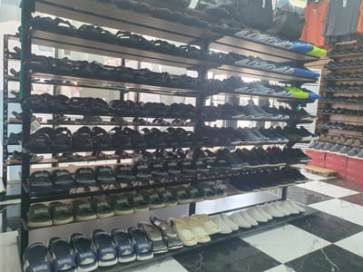 The image depicts the interior of a shoe store located in Suzak, Kyrgyzstan. The store features multiple shelves stacked with various types of footwear, primarily sandals and athletic shoes. The shelves are made of dark wood and metal, and the store appears well-organized, with shoes neatly arranged. On the upper shelves, there are men's sandals in black and brown. Below these, more sandals are displayed, including a row of black sandals with the brand name "Caddeem" visible. Further down, green "Caddeem" sandals are arranged, followed by a row of blue and white sandals, also bearing the "Caddeem" logo. The lower shelves showcase a variety of athletic shoes in different colors, including blue, green, and grey. Some shelves also display white flat shoes. In the background, athletic apparel such as jerseys are visible hanging on a rack. The flooring is a distinctive checkered pattern of black and white marble tiles. No people are visible in the frame.