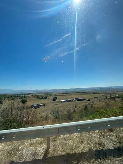 A wide, sun-drenched, panoramic view of a rural landscape in Gradets, Bulgaria. The sky is a vibrant, clear blue, with the bright sun casting a lens flare that creates diagonal light streaks and numerous small circular reflections across the upper portion of the image. wisps of white clouds are visible. The foreground features a silver metal guardrail, likely part of a road, with visible mounting holes and a red reflective strip on its right side. Beyond the guardrail, the terrain is dry and brown, with sparse, dry vegetation and scattered bushes. Further into the midground, a few small, weathered buildings or sheds with corrugated metal roofs are visible among fields of dry grass and sparse trees. Some of these structures appear to be agricultural outbuildings. In the background, rolling hills fade into a distant, hazy mountain range under the bright sky. The overall impression is one of an open, arid, and sparsely populated rural area under a clear, sunny day.