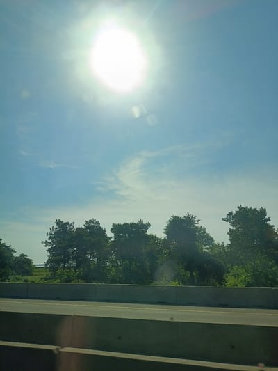 A bright sun shines through a clear blue sky with a few wispy clouds. In the foreground, a concrete barrier separates a road from a grassy embankment. Trees line the top of the embankment. The image appears to be taken from a moving vehicle. The location is not specified in Tlustenkhabl, Russia.