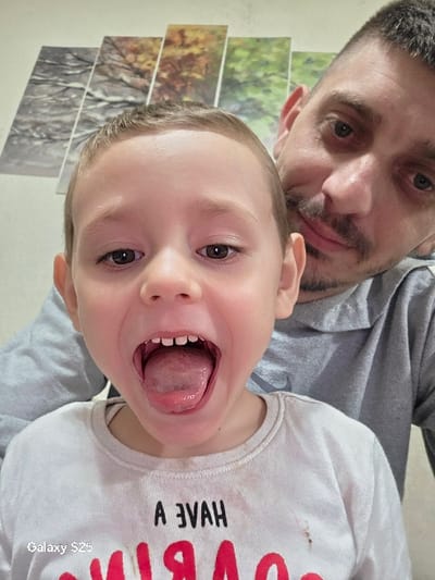 In the city of Karnalovo, Bulgaria, an indoor close-up portrait depicts a young child and an adult male. The child, with light brown hair, occupies the foreground, facing the camera directly. Their mouth is wide open, tongue extended, revealing several upper and lower teeth. The child wears a white long-sleeved shirt with partially visible red lettering on the front, including the sequence "A EVAN" and a large "R". Behind the child and to their right, an adult male with short dark hair and stubble is partially visible, looking towards the camera with a subtle expression. He is dressed in a grey hooded sweatshirt. The background consists of a light-colored wall adorned with four horizontally arranged rectangular canvas prints, each depicting a distinct seasonal tree landscape, from left to right: winter, autumn, and two scenes featuring green foliage. The image contains a "Galaxy S25" watermark in the lower left corner.