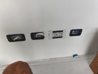 In this image, the scene is an indoor setting, likely a wall under construction or renovation, in Padua, Italy. The primary focus is on electrical work in progress. Three electrical boxes are visible. The leftmost box contains white electrical wiring. The middle box features a white double electrical socket that is partially installed, with more wiring visible behind it. The rightmost box shows some blue and white coiled wire.

The wall itself is painted white, and the lower portion of the image shows the base of the wall where it meets a ribbed surface, possibly a floor or a plinth. There is some tape or protective material along the base of the wall, with some peeling away, indicating an ongoing project. A roll of blue and white coiled material, possibly tape or conduit, rests on this base. In the foreground, a person's head and shoulder, with visible hair, are present at the bottom left of the frame, suggesting someone is either taking the photo or is involved in the work. The lighting appears to be artificial and even, with no strong shadows, suggesting an indoor environment. No specific time of day or weather conditions are discernible. There is no visible text in the image. The overall impression is one of a construction or electrical installation process.