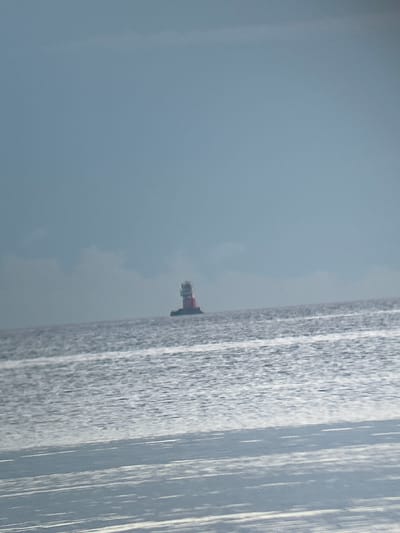 The image depicts a lighthouse in the sea, with a hazy sky and calm water. The lighthouse, with its red and white structure, is situated on a dark base. The water reflects the light, creating shimmering patterns. The overall atmosphere is serene and somewhat muted due to the overcast sky. The lighthouse is a navigational aid.