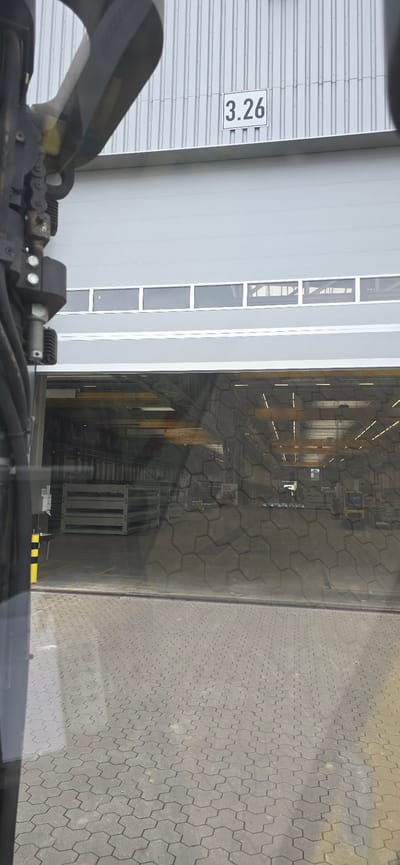This is an outdoor, daytime image taken from a vehicle, likely a forklift or similar machinery, looking towards a large industrial building with a metal exterior. The foreground on the left is a gray, interlocking paver surface. Through a large window or opening on the left side of the building, an industrial interior is visible, filled with metal racks, yellow beams, and what appears to be some sort of manufacturing or storage equipment. The floor of the interior also looks like it's made of interlocking pavers.
On the right side of the image, the building has a corrugated metal facade. There are several tall, narrow windows with white frames. A white sign with the black text "3.26" is attached to the corrugated metal on the right side of the building. The overall scene suggests a logistics or manufacturing facility. There are no people visible. The lighting indicates it is daytime, and the weather appears to be fair. The location is identified as Haren (Ems), Germany.