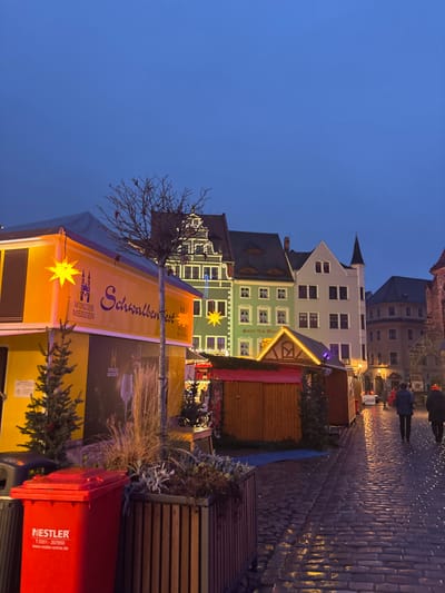 A European town square at dusk features cobblestone streets and historic buildings. A Christmas market is set up with stalls decorated with lights and signage for "Schallbenfest." A red NESTLER trash bin is visible in the foreground, along with a small fir tree. A few people walk in the distance.