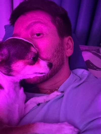 A close-up selfie shows a man with short dark hair and stubble lying down, with a small dog nuzzling his face. The image is illuminated with pink and purple light, possibly from a LED lamp or colored lighting. The man is wearing a light blue t-shirt. The dog has white and brown markings. In the background, a dark blue pillow and a patterned blanket are visible, suggesting the scene takes place indoors on a bed or couch. There is no visible indication of the location being in the city of Tokaryovo, Russia.
