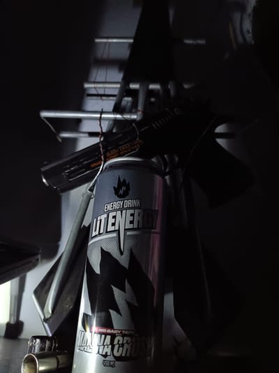 In a dimly lit indoor environment, possibly a workshop or storage area, a black Desert Eagle pistol is placed on a metal rack. Beside the pistol, a can of "Lit Energy" drink with a "Banana Crush" flavor is also positioned on the rack. The can is silver and grey with black graphics, and the pistol has orange lettering that reads "ISRAEL MILITARY INDUSTRIES". The scene appears to be a still life arrangement with the objects sharply in focus against a dark, indistinct background. The location within Tash-Dobo, Kyrgyzstan, is not evident from the visual information provided.