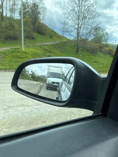 The image depicts a side view mirror of a vehicle, reflecting a white van on a road. The background outside the mirror shows a grassy hill and a tree with bare branches. The interior of the vehicle is visible on the right side of the frame, showing a gray surface. It is difficult to verify that this photo was taken in Верхняя Мара, Russia, as the location cannot be determined from the image alone.