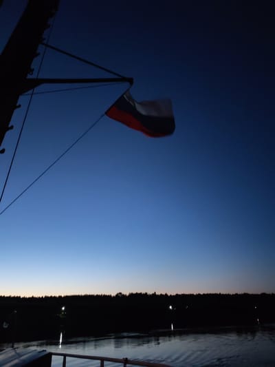 The image depicts a dark blue twilight sky over a dark, silhouetted treeline bordering a body of water. A Russian flag is visible, fluttering from a mast on the left side of the frame, which is part of a dark, indistinct structure. The water reflects the faint lights from the treeline. The scene appears to be a natural landscape at dusk, possibly near a body of water in a rural or forested area.