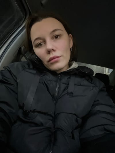 The image is a close-up, rotated selfie featuring a young individual, likely female, with dark brown hair. She has well-groomed dark eyebrows and brown eyes, and is looking directly at the camera with a neutral to slightly soft expression. A small, thin silver hoop earring is visible in her left ear. She is wearing a bulky, black puffer jacket with a prominent black zipper down the front and at least one black strap or buckle detail on the chest area. A light-colored, possibly white or light grey, fabric is visible around her neck, suggesting an undershirt or hoodie. The background is largely dark and indistinct, with some faint glints and shadows that suggest an indoor or vehicle setting. In the bottom left portion of the image, a metallic or light-colored frame, consistent with a car door or window interior, is visible. The lighting is dim, highlighting the subject's face against the dark surroundings. No text is visible in the image.