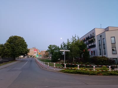 The image depicts an outdoor scene in Grójec, Poland, during what appears to be dusk or early evening, as indicated by the soft, pale sky and the presence of a visible moon. The setting is a street, with a road curving to the left, bordered by a sidewalk and a red and white striped barrier. On the right side of the road, a modern, multi-story apartment building with balconies is prominent. Further down the street, more buildings and trees are visible. On the left side of the road, a large, mature tree dominates the foreground.
Several cars are present on the road, and a pedestrian crossing is marked. Traffic lights are visible, with one showing a green light. There is a street signpost with two signs, one reading "ul. Mszczonowska" in Polish.
Notable details include the clear, unblemished sky, suggesting fair weather. The overall mood is calm and peaceful. There are no people clearly visible in the image. The scene appears to be a typical urban or suburban street at the end of the day. Visible text includes the street name "ul. Mszczonowska" and some signage on buildings in the distance, which are not legible enough to decipher specific details. There is also a small red sign on the apartment building.
