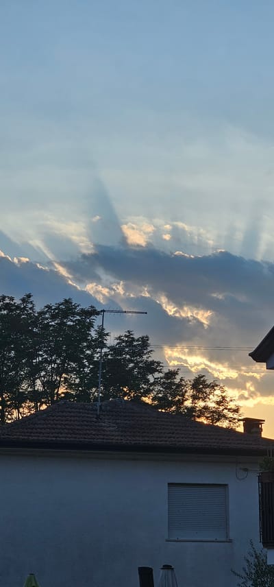 The image captures a sunset scene over a residential area in Villa Latina, Italy. The sky is a blend of soft blues and whites, with dramatic light rays piercing through scattered, dark clouds, illuminating patches of golden light. Silhouetted against the sky are the tops of lush green trees, and a television antenna. In the foreground, the tiled roof of a house is visible, with a white wall and a window covered by a closed, slatted blind. To the right, a portion of another house's roof and wall is visible, along with a chimney. The overall impression is one of a quiet, end-of-day atmosphere in a suburban setting.