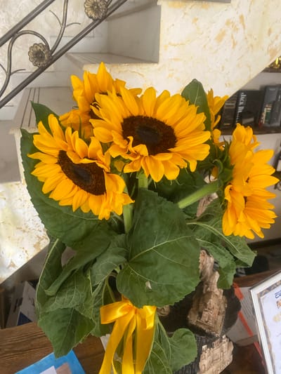 A bouquet of sunflowers with large green leaves is displayed in a vase. A yellow ribbon is tied around the stems. In the background, there is a staircase with a decorative railing, and a bookshelf filled with books. The scene is indoors, possibly in a residential setting.