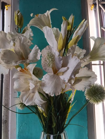 A close-up, vertical shot features a bouquet of white artificial lilies and green foliage in a clear glass vase. The flowers have delicate, ruffled petals and some exhibit subtle green speckling near the center. Several unopened buds, tinged with yellow, are also visible among the blooms. In the foreground, to the right, a spiky, green spherical accent piece is positioned. The background is a vibrant teal-colored wall, with a partial view of a window frame and blinds on the right side. The lighting suggests an indoor setting, possibly near a window, with natural light illuminating the scene. The image appears to be taken in Cibungbulang, Indonesia, based on the provided information. There are no people or animals present in the image.