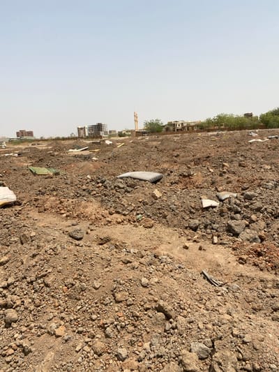 The image shows a dirt field in Khartoum, Sudan, at the coordinates 15.528 latitude and 32.571 longitude. The field is covered with rocks and rubble, and there is a building in the distance. There are several mattresses scattered throughout the field. The sky is clear and blue. There is a mosque minaret in the distance. There are no people in the image.  The location appears to be an abandoned area.  It is unclear if the mattresses were intentionally left behind.  The area looks like a dumping ground for mattresses and construction waste.