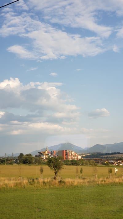 The image shows a panoramic view of the landscape surrounding the Gino Paradise Bešeňová water park in Bešeňová, Slovakia. In the mid-ground, the distinct colorful structures of the water park are visible, including slides and associated buildings. Behind the water park, a range of mountains forms the backdrop, partly obscured by clouds. The foreground consists of green fields and some scattered trees. The sky is blue with scattered white clouds. The scene appears to be a sunny day. There are no people or animals clearly visible in the image.