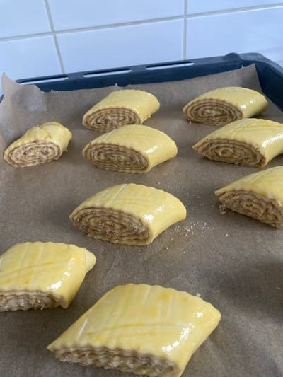 The scene depicts nine unbaked pastries arranged on a light brown parchment-lined dark baking tray, observed from an elevated perspective. Each pastry is roughly rectangular with rounded edges, coated in a glossy, pale yellow egg wash, and features three parallel score marks across its top surface. The cut ends of the pastries reveal concentric layers of light-colored dough enclosing a lighter brown, granular filling. Scattered white crystalline particles, possibly sugar or salt, are visible on the parchment paper around several pastries. The dark baking tray features a raised rim. In the upper background, a white tiled wall with dark gray grout lines is visible, indicating an indoor kitchen environment. The specific location within Chantepie, France, cannot be determined from the visual information presented.