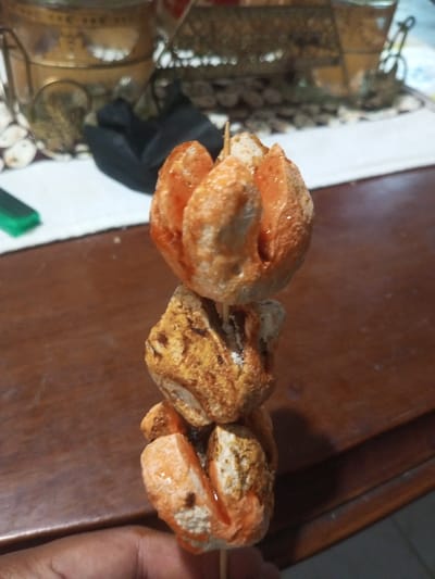 A close-up shows a skewered snack. The food, possibly fried fish balls or tofu, is stacked vertically on a wooden stick. The top and bottom pieces are coated in an orange glaze, while the middle one appears lightly browned. The snack is held near a dark wood surface. In the blurred background are decorative metal objects on a patterned surface.