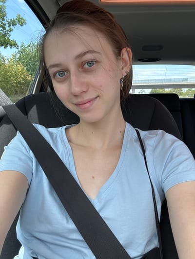 A young woman with light blue eyes and brown hair pulled back is sitting in the driver's seat of a car, wearing a light blue V-neck t-shirt. She is looking at the camera with a slight smile. A black seatbelt is across her chest. The background shows trees and a blue sky visible through the car window, suggesting an outdoor setting. The scene is consistent with being in or near Großwallstadt, Germany, a town known for its natural surroundings.
