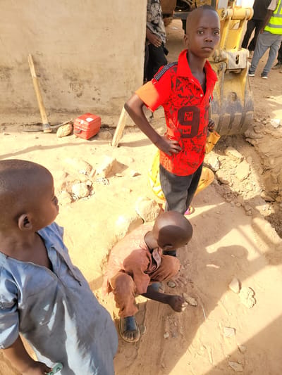 The image depicts an outdoor scene in Nassarawa Egon, Nigeria, characterized by dry, light brown soil and an apparent construction or excavation site.

In the foreground, three children are present. A young male, approximately 8-10 years old, stands centrally, facing the viewer with a neutral expression. He wears a short-sleeved red collared shirt with black abstract patterns and a prominent black number "9" on the front, along with dark trousers. His hands are positioned on his hips. To his left, another young male, of similar age, is partially visible from the chest up, facing right. He wears a light blue, possibly soiled, long-sleeved garment and holds a small, light green object in his right hand. To the right and slightly in front of the central child, a third young male, likely younger than the other two, crouches on the ground. He wears a light brown tunic and dark sandals, focusing his attention on small rocks and debris on the ground, manipulating them with his hands near a slender white object.

The background reveals a construction environment. A large yellow excavator bucket is prominently positioned to the right of the central child, partially submerged in an excavated trench. Behind the excavator, several adult figures are partially visible; one wears grey trousers and a yellow high-visibility vest, another wears dark attire. To the left, a light-colored, possibly mud or concrete, wall stands, against which a wooden-handled pickaxe is leaned. On the ground near the wall, a red plastic container with a white label and a woven, natural fiber object are present. The ground throughout the scene is marked by small rocks, dirt, and varied textures indicative of excavation. Strong shadows cast on the ground suggest bright daylight.