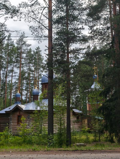 The image shows a small wooden church with blue domes and golden crosses, situated in a forest near Nenimyak, Russia. The church is made of logs and has a metal roof. In front of the church, there is a dirt road with scattered pinecones. The trees surrounding the church are tall and dense, typical of a Russian forest. The sky is overcast, suggesting a cloudy day. There are no people visible in the image. The scene is peaceful and appears to be in a rural or remote area.
