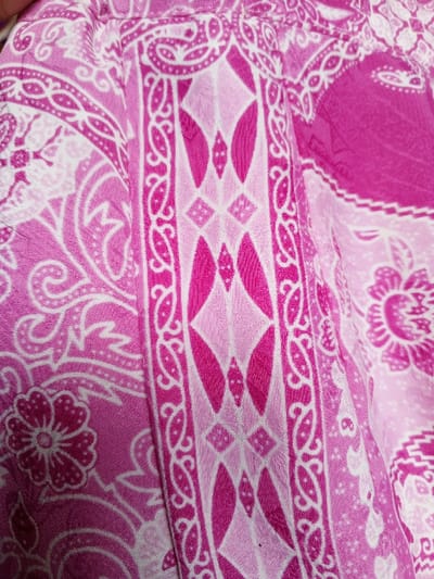 The image showcases a close-up of patterned fabric, primarily in varying shades of pink and white. Intricate designs fill the space, including paisley motifs, floral patterns, and a central vertical stripe featuring diamond shapes and decorative borders. The fabric appears textured, with subtle tonal variations within each colored section, suggesting a woven or printed material. The overall impression is of a vibrant and detailed textile design.