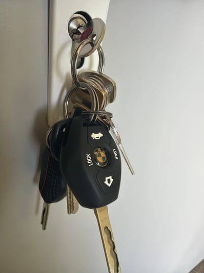 A close-up, slightly angled shot captures a set of keys hanging from what appears to be a key rack or hook mounted on a white, vertical surface. The keys are on a silver-colored keyring. The most prominent key fob is black, featuring the BMW logo in its center, which has a yellow and blue circular design. The letters "LOCK" are printed in white text below the BMW logo. Above the logo, a white silhouette of a car trunk is visible. Another key, a car key with a black textured grip and a red stripe, hangs to the left of the BMW key. Two other metallic keys are also present, one with a distinct golden color and an intricately cut blade. The white surface behind the keys is a plain, neutral background. The scene is devoid of any people or any indication of a specific location within Dinkata, Bulgaria. The lighting suggests an indoor environment, possibly a home or office, with the light source casting minimal shadows.