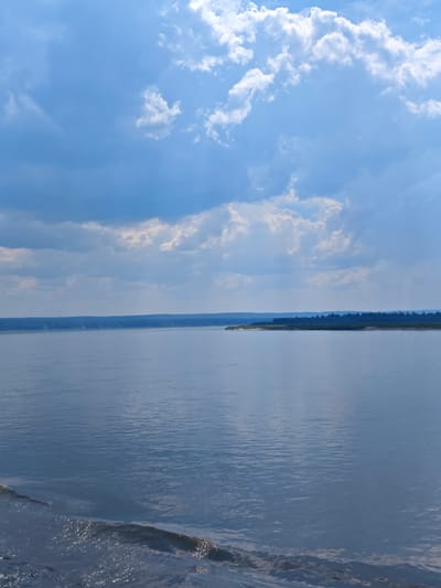 The image depicts a wide, calm body of water, likely a river, with a densely forested shoreline in the distance. The sky above is a mix of bright blue and dramatic clouds, with sunlight breaking through in places. The water's surface reflects the sky, creating a serene and expansive atmosphere. The foreground shows a gentle wave breaking, suggesting movement in the water. Based on the location provided, this scene is set near Podkamennaya Tunguska, Russia, a region known for its vast wilderness and river systems. There are no people or explicit objects of human construction visible in the image, emphasizing the natural and untamed character of the environment. The scene is still and peaceful, with the primary elements being water, land, and sky.