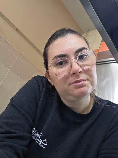 A close-up, slightly low-angle photograph captures an adult female within an indoor setting in Gevgelija, North Macedonia. The subject, positioned center-frame, has dark hair pulled back and wears thin, gold-rimmed, hexagonal glasses. Her expression is neutral. She is dressed in a black long-sleeved crew-neck sweatshirt featuring a white stylized text and coffee cup logo on the left chest. The background on the left consists of light beige wall tiles with a visible horizontal conduit. To the upper right, dark surfaces and a metallic appliance are partially visible, suggesting a commercial or kitchen environment.