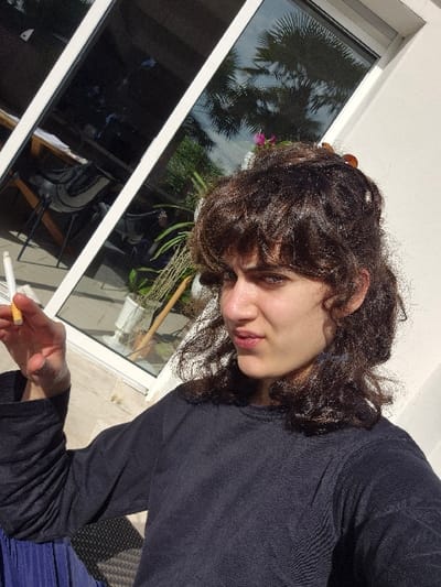 A young adult individual is depicted in a medium close-up, seated outdoors on a paved patio or terrace at a residence in Châtelaillon-Plage, France. The subject, possessing dark, curly hair styled partially upward with two visible brown clips, wears a dark long-sleeved top and a blue pleated lower garment. The right hand is elevated, holding an unlit white cigarette with an orange filter between the index and middle fingers. The subject's gaze is directed towards the viewer, maintaining a neutral facial expression. The scene is brightly lit by direct sunlight, highlighting the subject's face and hair. The background features a building's white exterior wall and large white-framed glass door panels that reflect a clear blue sky, a prominent fan palm tree, and other green vegetation, including a hint of pink/purple flowers. Reflections also show interior elements such as a table and chairs within the building. A white planter is partially visible behind the subject. A segment of a dark, woven outdoor chair is discernible beneath the subject's left arm.