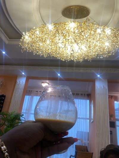 A person is holding a glass of an amber-colored liquid, possibly alcohol. The person's hand is dark-skinned. The background features a large, ornate chandelier with a multitude of crystal beads. The chandelier is hanging from a white ceiling with an intricate design. The room appears to be a living room or dining room, with large windows and white curtains, as well as a wall with a light peach color. The overall scene suggests a luxurious home in a residential area of Buea, Cameroon. The coordinates provided, latitude: 4.145, longitude: 9.23, correspond to a location in Buea, Cameroon.