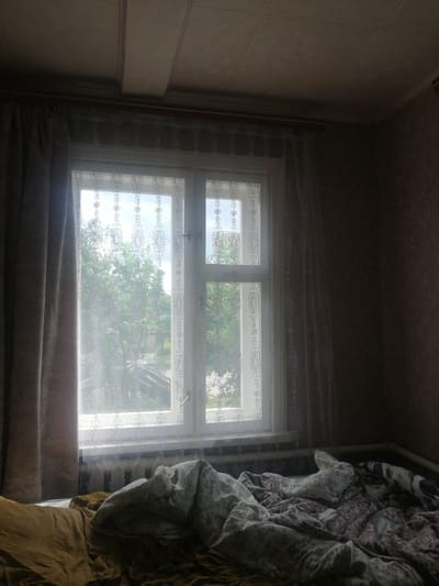 The image depicts an interior view of a bedroom in Dubrovka, Russia, looking towards a window. The window is partially covered by a thick, brown curtain on the left and a sheer, white lace curtain with a floral pattern on the right. The window itself is a double-paned unit with a white frame, and through it, lush green trees and a cloudy sky are visible. Below the window, a radiator is present. The foreground of the image shows a bed with rumpled bedding. The bedding is a mix of patterns and colors, including brown, gray, and white, with one pillow visible. The walls appear to be textured and wallpapered in a muted tone. The ceiling is paneled with visible seams. The lighting suggests it is daytime, with sunlight filtering through the window, creating some shadows and highlighting the dust on the lace curtain. No people are visible in the scene.
